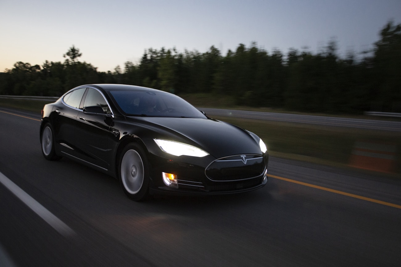 Чёрная Tesla Model S с включёнными фарами на трассе в сумерках